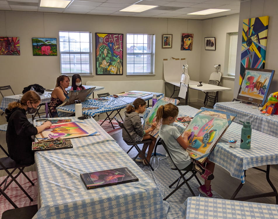 Children sit at tables with blue and white gingham tablecloths, using oil pastels to illustrate still-lifes