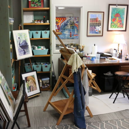 Art studio with an easel in the middle, aprons hanging off the side of the easel and different pieces of art are displayed around the room.