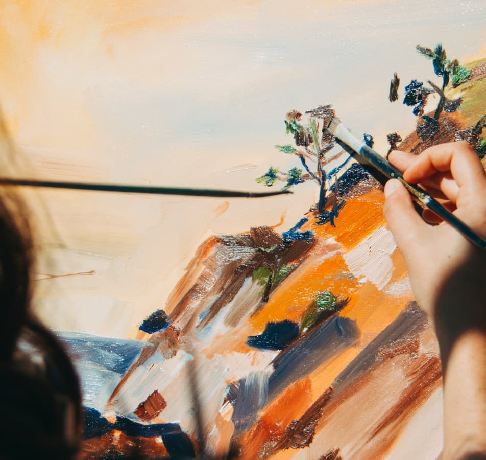 Close up photo of a hand holding a paintbrush, adding leaves on a tree on an impressionist style painting