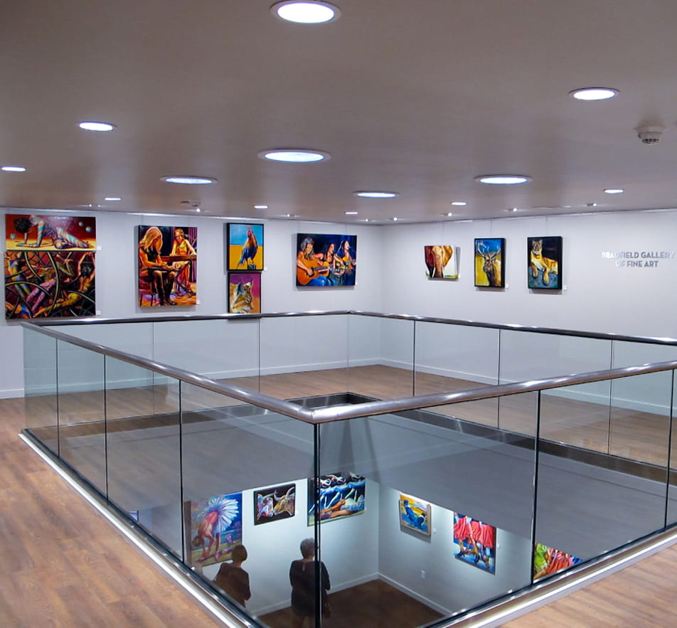 Second floor of an art gallery with a glass-fence balcony that looks down to the first floor. You can see paintings hanging on the walls of both floors.