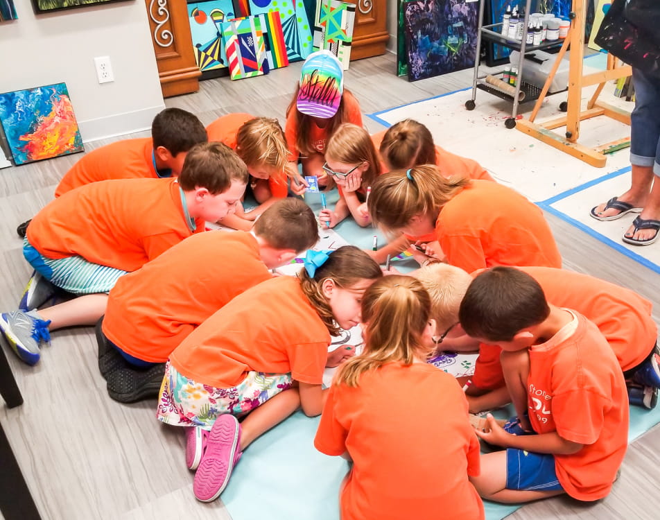 A group of children in bright orange shirts crouch on the ground, coloring on a large piece of paper.
