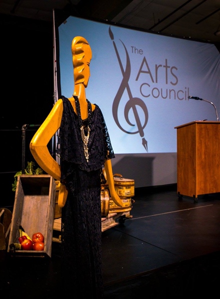 Stage with a podium and the Arts Council logo projected on the back wall. There's an abstract mannequin wearing a black lace dress and a gold and black necklace in the foreground.