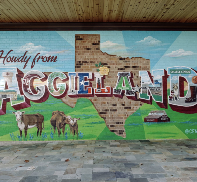 Mural of a green, neatly mowed pasture and blue skies on a brick wall. There is an unpainted area in the middle in the shape of Texas. Cows graze alongside bluebonnets and the words 'Howdy from Aggieland' are painted in block letters over top of it all.