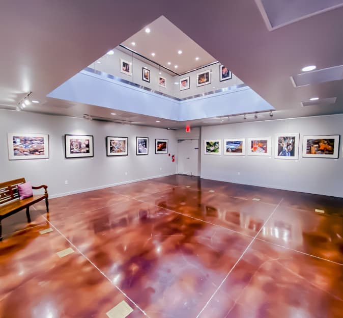 View from the first floor of an art gallery, you can see up to the second floor through a skylight. Artwork is hanging on the walls of both floors.