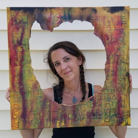 Headshot of Christine Holton holding a graffiti covered canvas. She is looking through the hole that has been ripped in the middle of the canvas.