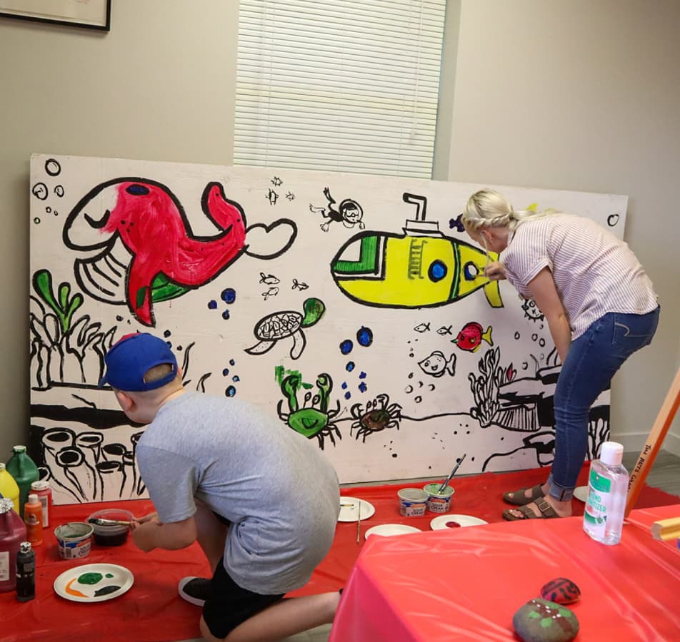 A woman and a young boy paint an underwater ocean scene on a large white canvas