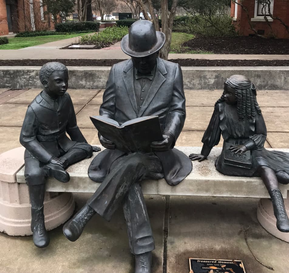 Metal statues of a man and two children sitting on a concrete bench, reading a book