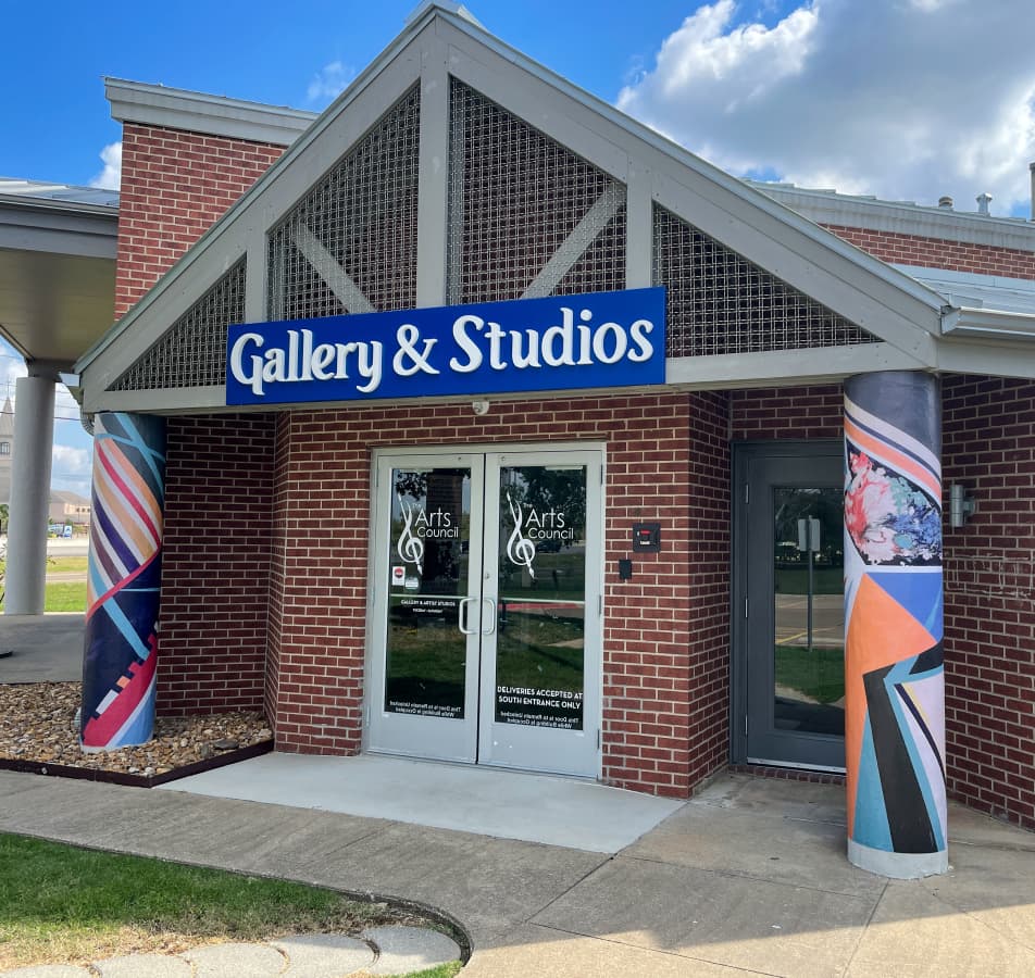 Brick building with a blue sign that says 'Gallery & Studios' in white letters on the front and colorfully painted pillars on either side of a double door