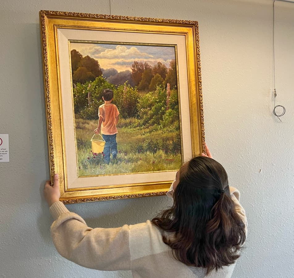 Woman hanging a gold framed painting of a little boy holding a yellow bucket in a meadow.