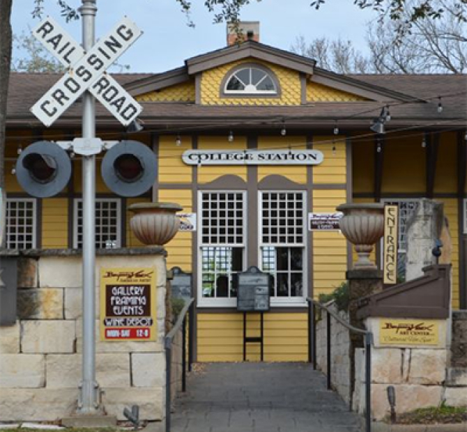 Refurbished yellow train depot with a railroad crossing sign in the front and a sign that reads 'College Station' above two grid pane windows.