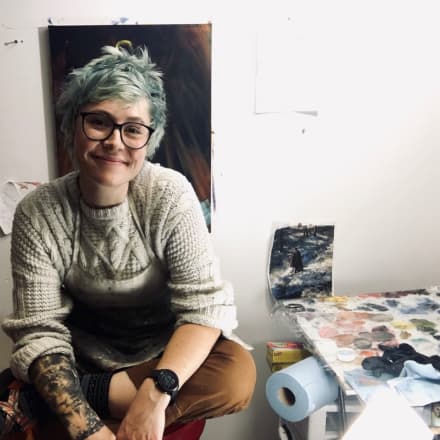 Saskia Becker sits cross legged on a stool next to a paint covered table.