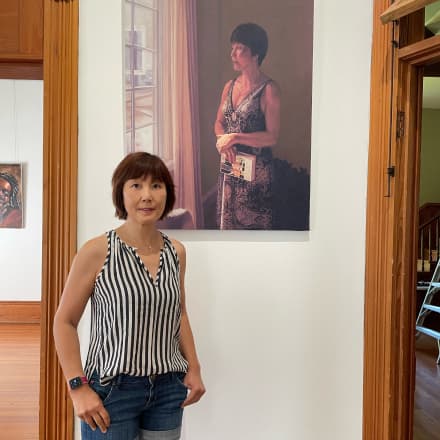 Siana Smith stands in front of a self portrait painting of her looking out a widow and holding a book