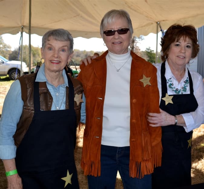 Three older women stand together smiling, the two on the ends are wearing black aprons and the woman in the middle is wearing an orange, suede fringe jacket and sunglasses.